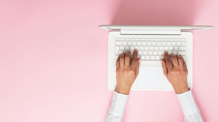 A top-down view of a woman's hands on a white laptop, against a pink background.