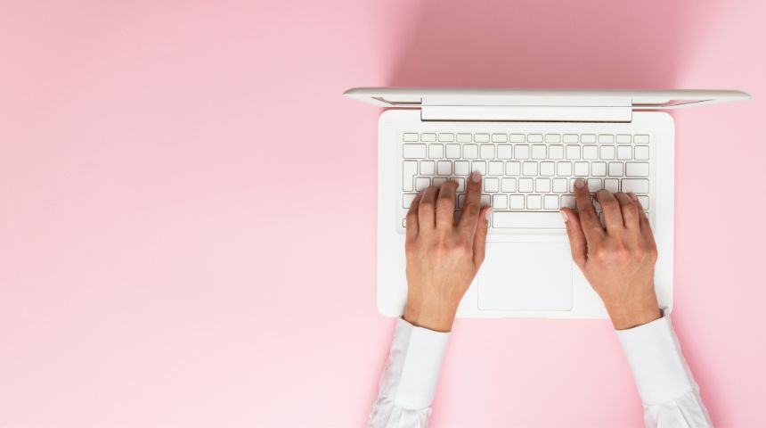 A top-down view of a woman's hands on a white laptop, against a pink background.