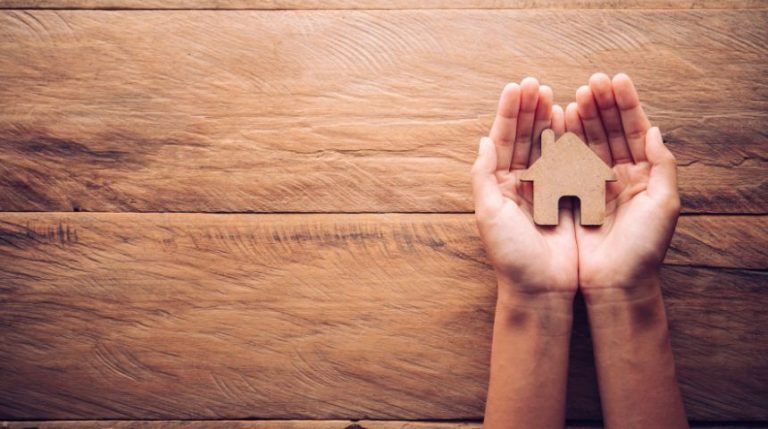 Two hands holding a small wooden model of a house.