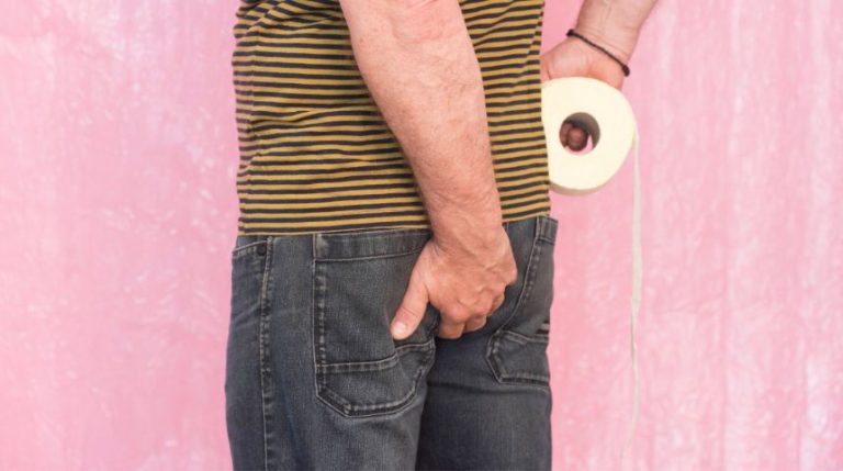 A man with one hand on his backside, and the other holding a roll of toilet paper.