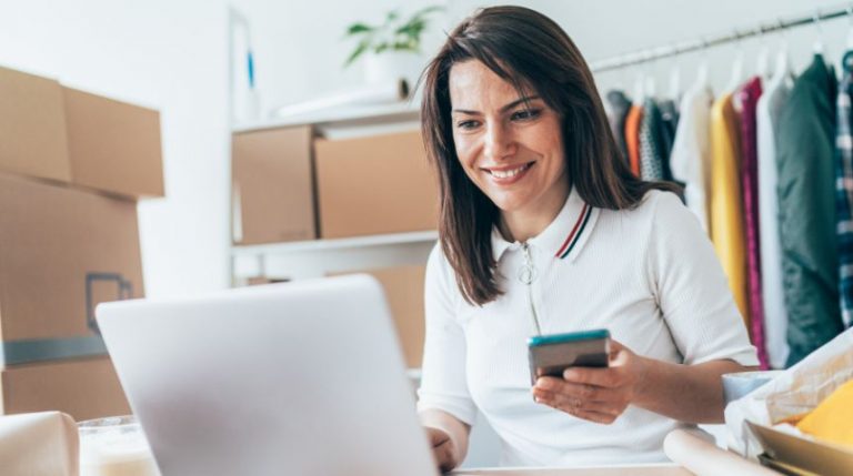 A woman sitting at a desk with her laptop and cell phone. She's surrounded by boxes and stock.
